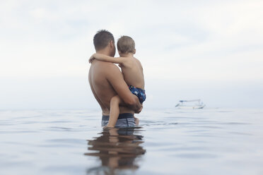 Side view of shirtless father carrying son while standing in sea against cloudy sky - CAVF54150