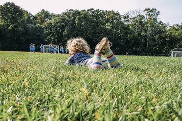 Girl looking at soccer match while lying on grassy field at playground - CAVF54191