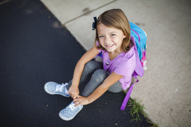 High angle portrait of smiling girl with backpack sitting on footpath - CAVF54226