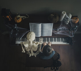 Hoher Blickwinkel des Jungen, der Klavier spielt, während er neben einem Skelett sitzt, während Halloween - CAVF54232