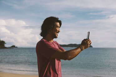 Seitenansicht eines Mannes, der mit seinem Mobiltelefon am Strand gegen den bewölkten Himmel fotografiert - CAVF54237