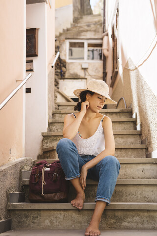 Nachdenklicher Tourist mit Hut, der mit seinem Rucksack auf einer Treppe in der Stadt sitzt, lizenzfreies Stockfoto