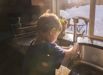 Junge wäscht sich im Winter die Hände an der Küchenspüle am Fenster - CAVF54323