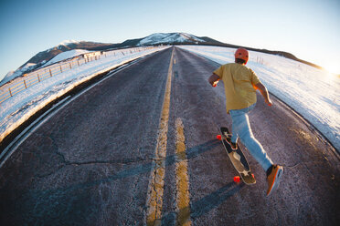 Rear view of man skateboarding on road amidst snow covered field against clear sky - CAVF54329