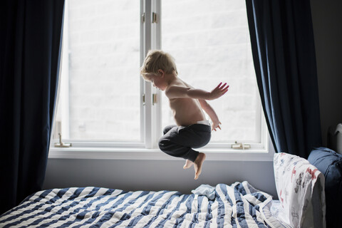 Hemdloser Junge, der zu Hause auf dem Bett am Fenster springt, lizenzfreies Stockfoto