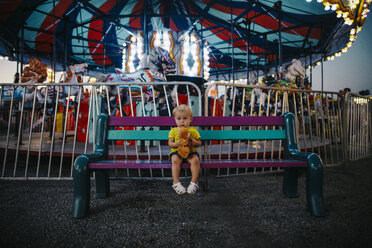 Portrait of baby girl holding toy while sitting on bench at amusement park - CAVF54358