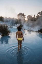 Rückansicht einer Frau, die einen Bikini trägt, während sie bei Sonnenuntergang im Thermalbad spazieren geht - CAVF54384