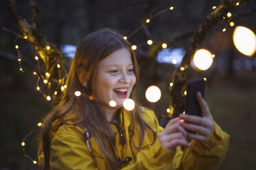 Fröhliches Mädchen mit Smartphone inmitten von beleuchteten Lichterketten Dekoration in der Nacht - CAVF54390