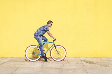 Side view of man riding bicycle against yellow wall at sidewalk in city - CAVF54408