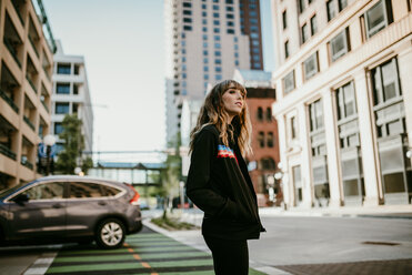 Side view of young woman with hands in pockets standing on city street - CAVF54419