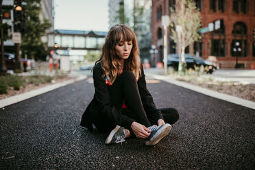 Young woman tying shoelace while sitting on city street - CAVF54421