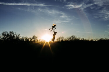 Playful silhouette father throwing daughter in air while playing with her on field against sky during sunset - CAVF54490