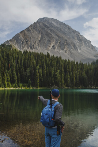 Rückansicht eines Wanderers mit Rucksack, der in Richtung See zeigt, während er vor Bergen im Wald steht, lizenzfreies Stockfoto