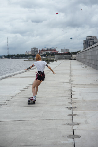 Rückansicht einer jungen Frau auf einem Carver-Skateboard am Flussufer, lizenzfreies Stockfoto