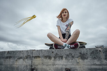 Young woman sitting on a concrete wall on carver skateboard with kite in background - VPIF00997