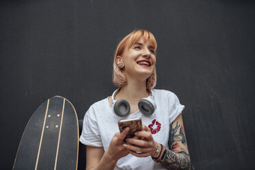 Glückliche junge Frau mit Skateboard, Kopfhörern und Mobiltelefon - VPIF01017