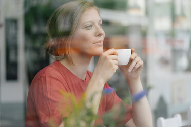 Young woman with cup of coffee behind windowpane in a cafe - KNSF05310