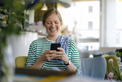 Lachende junge Frau, die in einem Café auf ihr Handy schaut - KNSF05353