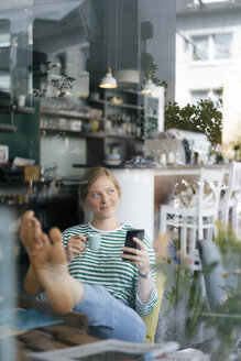 Lächelnde junge Frau mit hochgelegten Füßen, die ein Mobiltelefon und eine Espressotasse in einem Café hält - KNSF05357