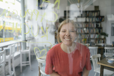 Porträt einer glücklichen jungen Frau, die hinter der Fensterscheibe eines Cafés posiert - KNSF05377