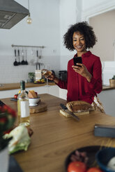 Woman drinking white wine in her kitchen, using smartphone - BOYF00971