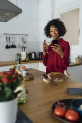 Woman drinking white wine in her kitchen, using smartphone - BOYF00972