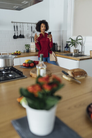 Frau trinkt Weißwein in ihrer Küche und benutzt ihr Smartphone, lizenzfreies Stockfoto