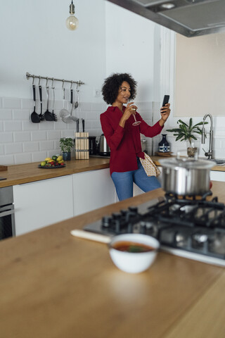 Frau trinkt Weißwein in ihrer Küche und benutzt ihr Smartphone, lizenzfreies Stockfoto