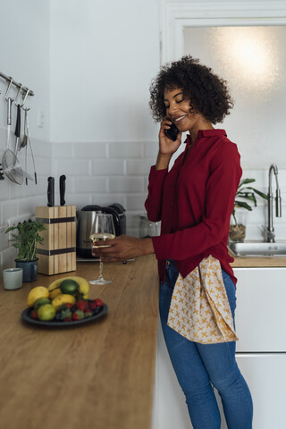 Frau steht in ihrer Küche, mit einem Glas Wein, und benutzt ihr Smartphone, lizenzfreies Stockfoto