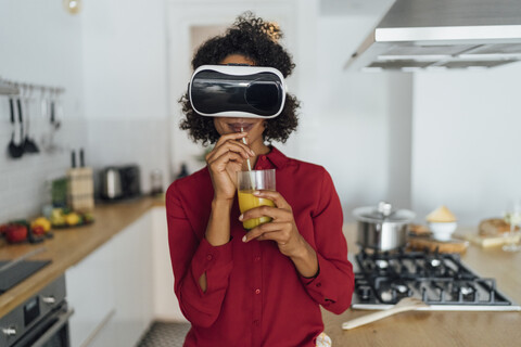 Frau steht in ihrer Küche, trägt eine VR-Brille und trinkt Orangensaft, lizenzfreies Stockfoto