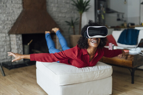 Frau liegt auf einem Sitzmöbel, trägt eine VR-Brille und tut so, als würde sie fliegen, lizenzfreies Stockfoto