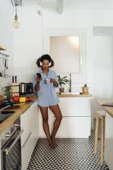 Frau mit Kopfhörern, die ihr Smartphone benutzt und Kaffee zum Frühstück in ihrer Küche trinkt - BOYF01031