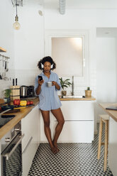 Woman with headphones, using smartphone and drinking coffee for breakfast in her kitchen - BOYF01031