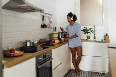 Woman with headphones, using smartphone and drinking coffee for breakfast in her kitchen - BOYF01033