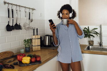 Woman with headphones, using smartphone and drinking coffee for breakfast in her kitchen - BOYF01034