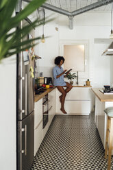Woman sitting on worktop of her kitchen, using digital tablet in the morning - BOYF01047