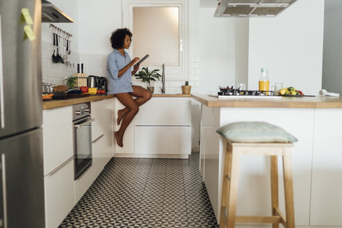 Woman sitting on worktop of her kitchen, using digital tablet in the morning - BOYF01050