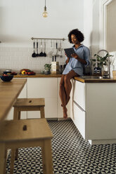 Woman sitting on worktop of her kitchen, using digital tablet in the morning - BOYF01052