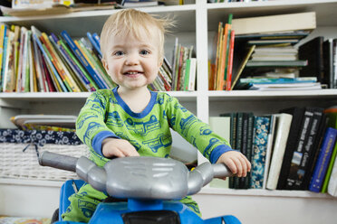 Portrait of baby boy riding toy motorcycle against bookshelves at home - CAVF54560