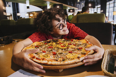 Happy woman holding pizza on wooden table while sitting in restaurant - CAVF54562