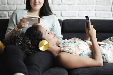 Woman using mobile phone while relaxing on girlfriend's lap reading book in living room - CAVF54575