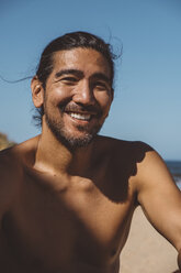 Portrait of shirtless man sitting at beach against clear blue sky during sunny day - CAVF54608