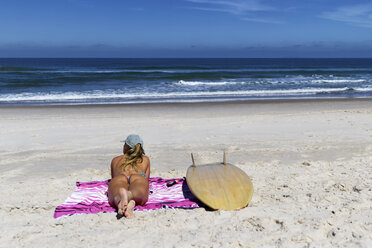 Rückansicht einer Frau im Bikini, die auf einem Surfbrett auf einer Decke am Strand liegt, gegen den blauen Himmel an einem sonnigen Tag - CAVF54609