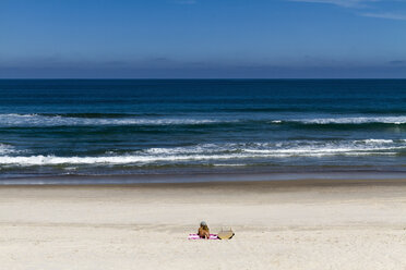 Mitteldistanzansicht einer Frau, die auf einer Decke am Strand neben einem Surfbrett liegt, gegen den blauen Himmel an einem sonnigen Tag - CAVF54610