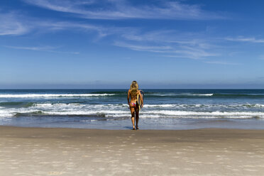 Rear view of woman in bikini carrying surfboard while walking towards sea against blue sky during sunny day - CAVF54611