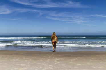 Rückansicht einer Frau im Bikini, die ein Surfbrett trägt, während sie gegen den blauen Himmel an einem sonnigen Tag zum Meer läuft - CAVF54611