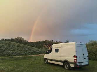 Glückliche Frau mit erhobenen Armen, die durch das Fenster eines Wohnmobils im Wald gegen den bewölkten Himmel bei Sonnenuntergang schaut - CAVF54616