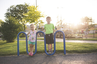 Ganzes Porträt des Bruders, der mit seiner Schwester auf einem gebogenen blauen Metall auf einem Spielplatz bei Sonnenuntergang steht - CAVF54627