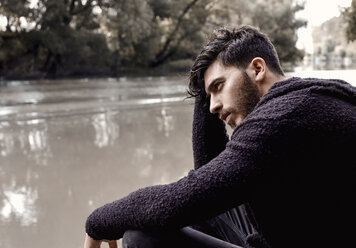 Thoughtful young man looking away while sitting by lake in forest - CAVF54653