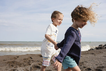 Happy siblings holding hands while walking at beach against sky during sunny day - CAVF54681